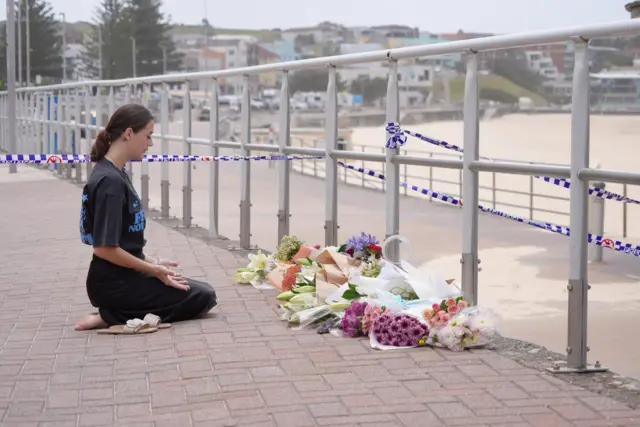 A woman kneels with her hands placed upwards and eyes closed in front of a makeshift flower memorial near metal railings and police tape with Bondi Beach visible in the background.