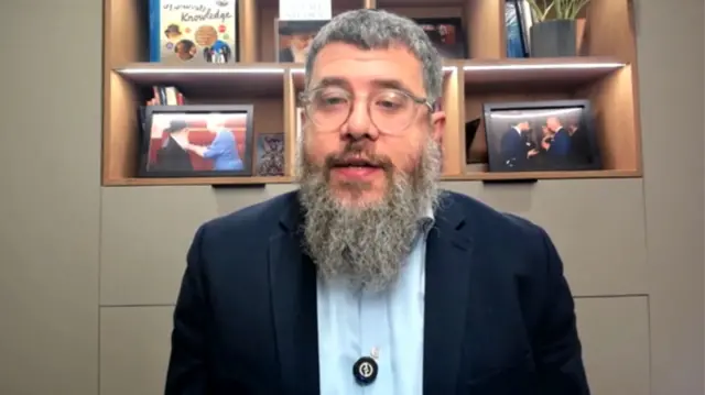 A man in a blue suit jacket and white shirt sits in front of book shelves