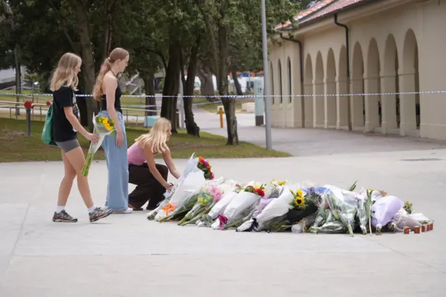 Three young women holding flowers stand in front of a makeshift memorial at Bondi Beach. One of the women kneel to place her bunch of flowers in a pile of other flowers.