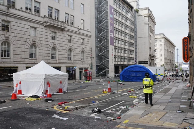 A police officer stands by a white tent with a large blue tent at the scene
