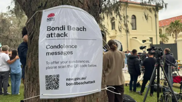 A poster has been fixed to a tree with details 'Bondi Beach attack'. Condolence messages. And a QR code