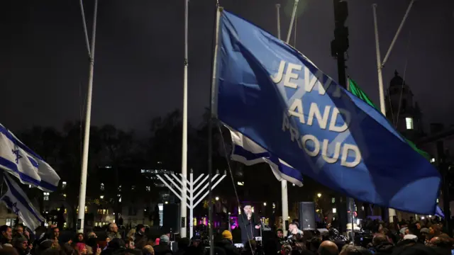 A large crowd at nighttime in London, a large blue flag flies over the crowd that reads "Jewish and proud"