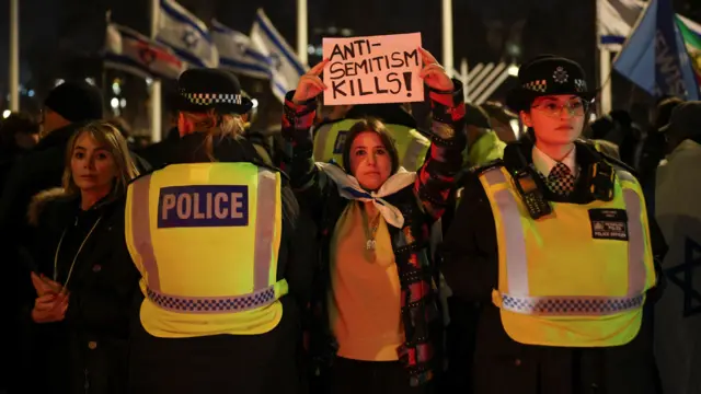 A woman holds up a sign that reads "anti-semitism kills", either side of her stand police officers watching the crowd