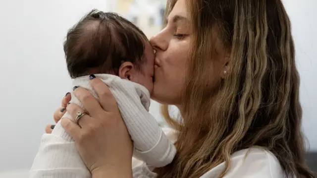 Chavi, a white woman with mousey brown hair, holding her baby and kissing their head, they are both wearing white.