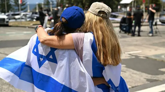 Two women embrace, both with the Israeli flag draped on their back