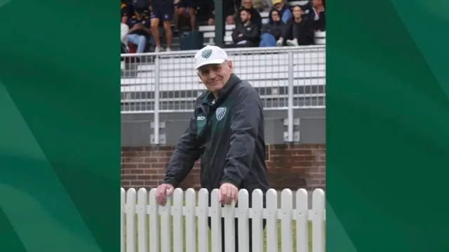 An elderly man in a black jacket and white cap smiling as he stands behind a white fence