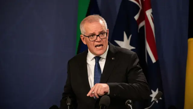 A man in a suit stands at a podium, an Australian flag is visible behind him