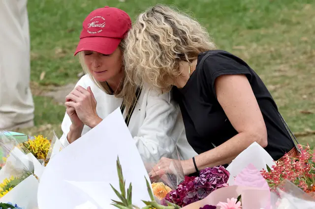 Two women, emotional, sitting next to large pile of flowers