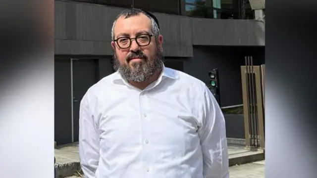 Rabbi Eli Schlanger stands with his arms crossed behind his back. he wears a white shirt and black glasses and has a thick grey beard