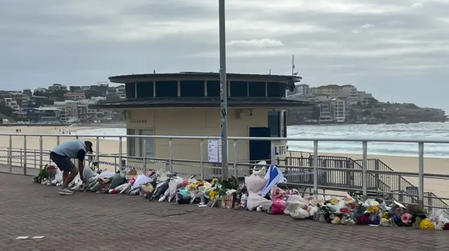 Bouquets of flowers are propped up against a railing along a promenade, with sand and blue waves visible behind them.