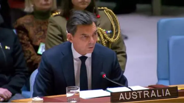 A man in a blue suit sits at a wooden table, a sign saying "Australia" is in front of him