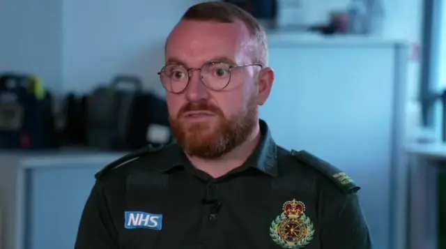 James Vernon, wearing his dark green paramedic's uniform, has short brown hair and beard. He wears dark, round glasses
