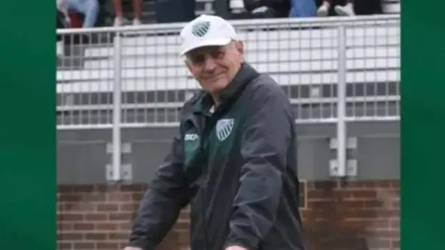 Peter Meagher stands at a white fence. he wears a rugby jacket and white cap