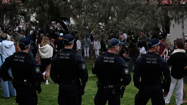 Four police guards stand with their back to the camera, watching the scene as a memorial gathers in a green patch on Bondi Pavilion