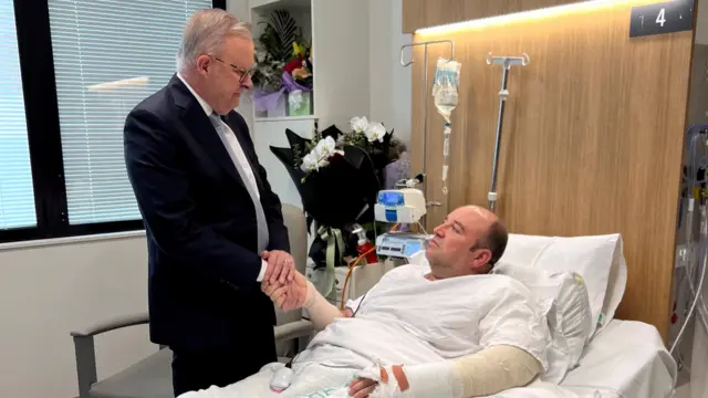 Australia's Prime Minister Anthony Albanese holds Ahmed al Ahmed's hand with both his hands. Al Ahmed lies in a hospital bed and his left arm is in a cast