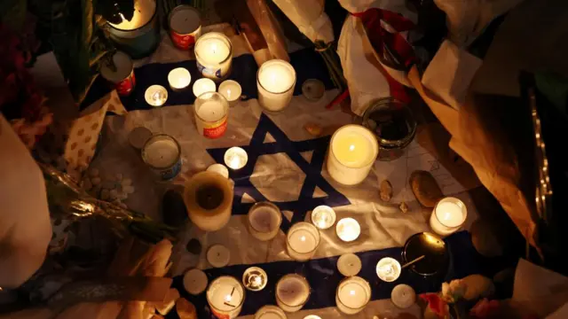 Dozens of lit candles sit on top of an Israeli flag. Several bunches of flowers have been placed next to the flag