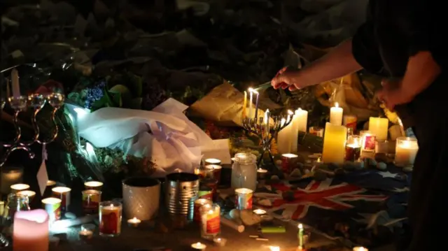 Many candles are lit on the ground - one person extends across the frame to light another