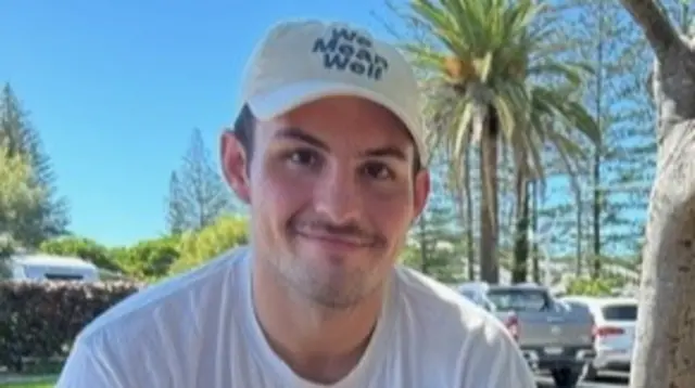 A man with a white T-shirt and a white baseball cap is pictured smiling.