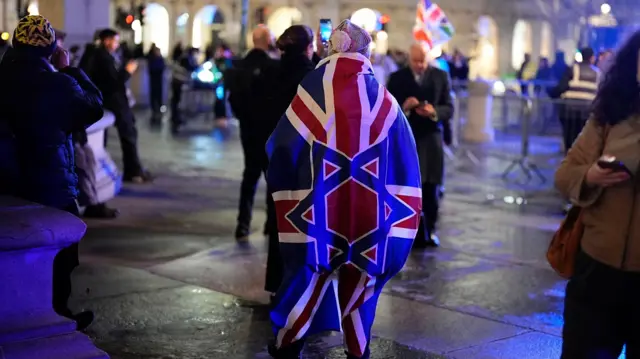 A person stands in Trafalgar Square, around their shoulders is a flag with the Union Jack and a star of David overlaid.