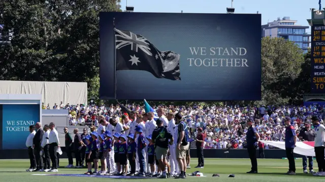 People are seen gathered on a cricket ground with heads bowed, beneath a billboard that reads "We stand together".
