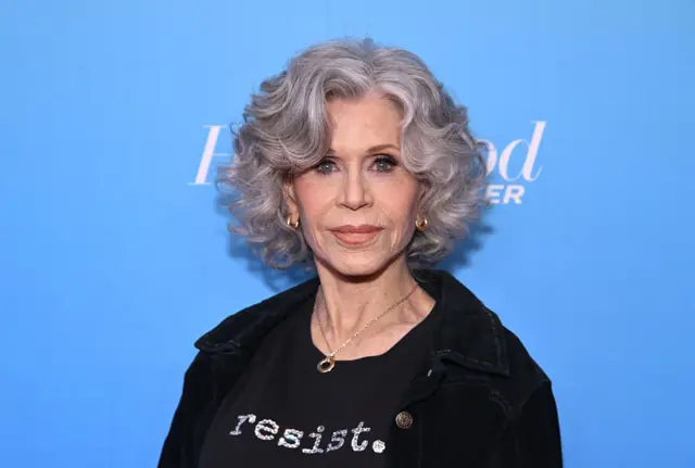 Jane Fonda stands in front of a blue backdrop. She is wearing a black jacket or a black shirt with the words 'resist' on the shirt.