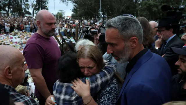 The mother of one of the victims of the attack hugs a woman in a blue checked shirt. Dozens of floral tributes can been behind the pair with a large crowd stood around looking at the tributes.