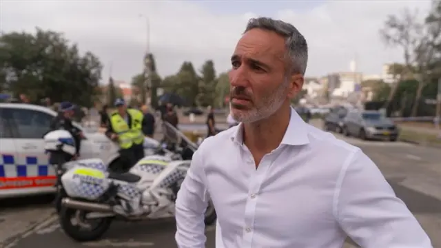A man wears a white shirt, in the background are a police car and motorbike.
