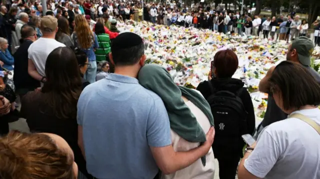 People stand around a mound of tributes - some hold each other