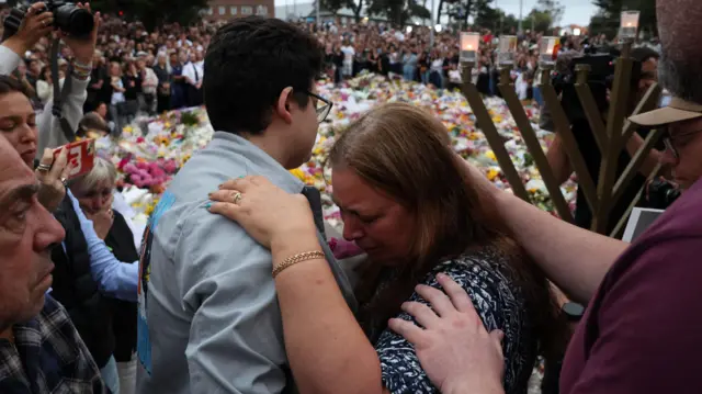 A crying woman with long brown hair hugs a person with short dark brown hair in front of a large crowd with flowers covering the floor between them