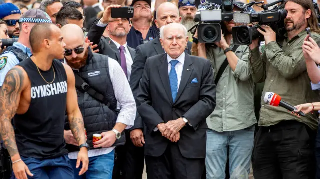 John Howard wears a suit and stands with his arms crossed. Members of the media stand beside him with cameras