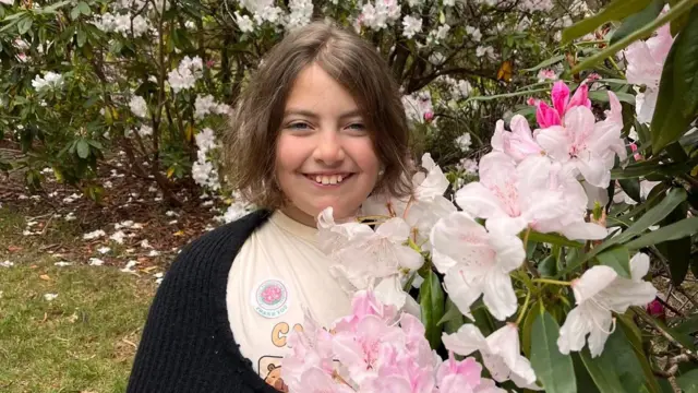 Matilda, a girl with short brown hair, smiles in a garden beside pink and white flowers.