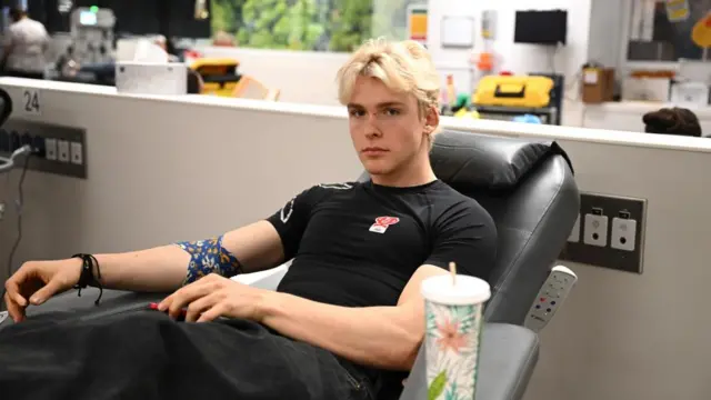 Tobias Bowgerald sits back in a black chair at a town hall. A brightly coloured cup decorated with flowers sits on the side of the chair