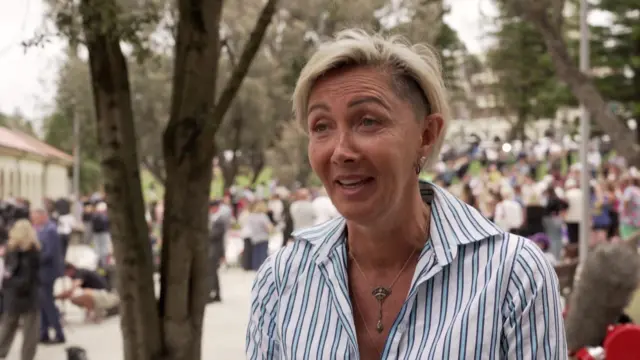 A woman wearing a blue and white striped shirt stands beside a tree with people in the background