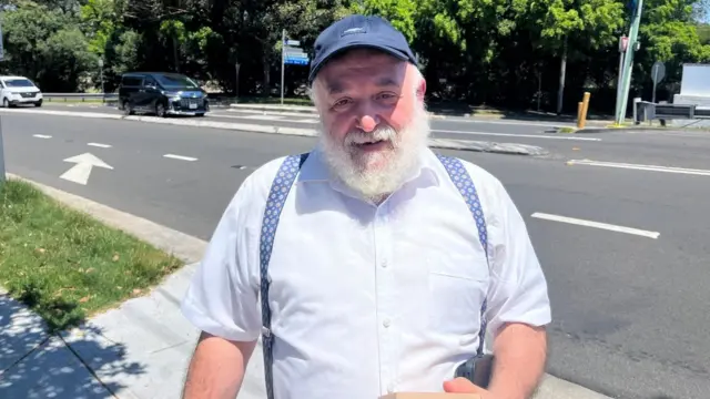 An older man with a white beard stood in the street in the sun. He's wearing a shirt and suspenders