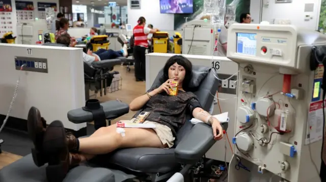 Mika Levy, 21, sits in a black chair with her legs crossed while drinking from a juice carton. Her left arm is out and has a line in it as she gives blood