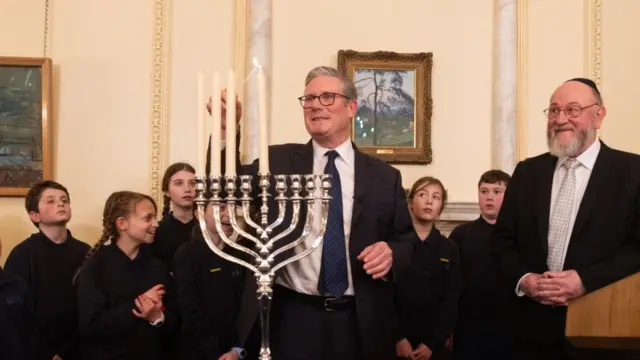 Starmer lighting a Jewish menorah in front of a group of children, and next to chief rabbi Sir Ephraim Mirvis