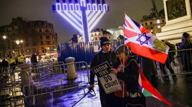 Two people stand in front of a giant lit-up Menorah, one of them holds a sign that says "British Jews do count" and has a flag bearing the Union Jack, Star of David and a lion emblem