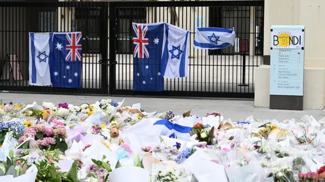 Australian and Israeli flags hang from a tall, metal gate. Across from the gate are rows of floral bouquets.