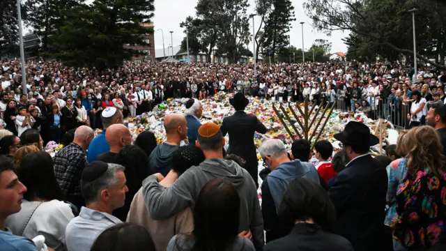 people gather in a large crowd in front of flowers and tributes on Bondi Beach