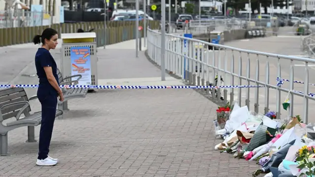 A woman in dark blue, short-sleeved scrubs and white trainers stands across from a metal railing lined with rows of floral bouquets