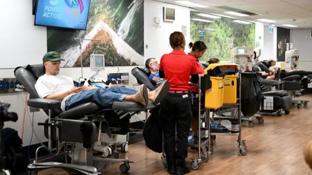 People lie down on reclined black chairs while medical staff work at trolleys