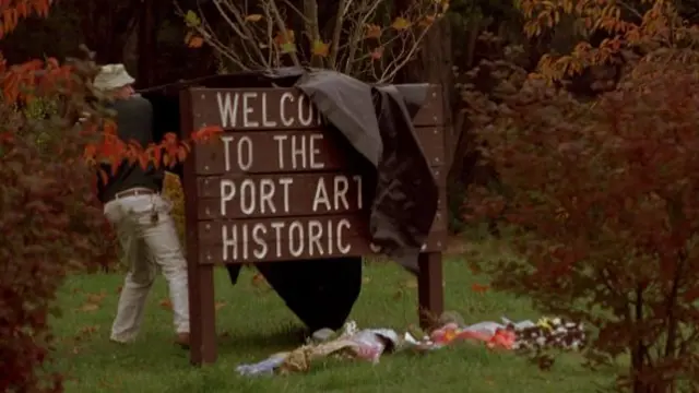 A man covers up a wooden sign with some tarpaulin, there are flower tributes laid out in front and the sign reads "Welcome to the Port Arthur Historic Site"