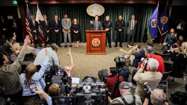 A crowd of journalists in a semi-circle face a line-up of Los Angeles law enforcement officials