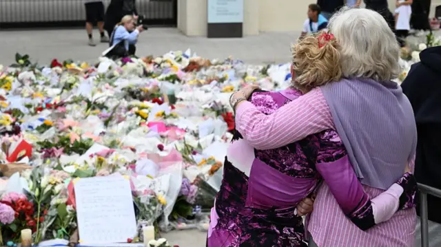 Backview of two women dressed in pink, hugging each other as they look at the flowers laying on the ground