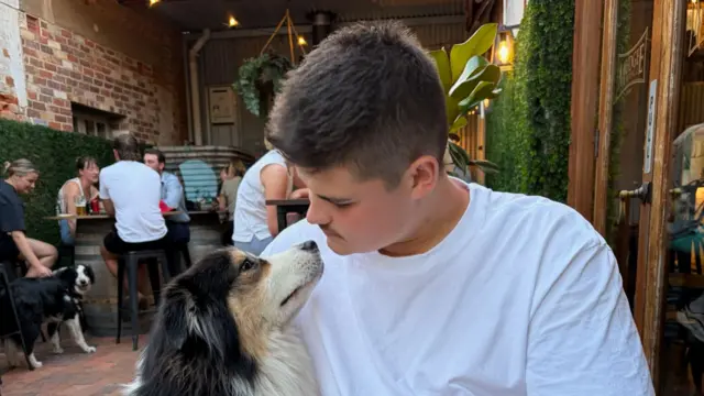 A young man in a white t-shirt is seen looking at a border collie.