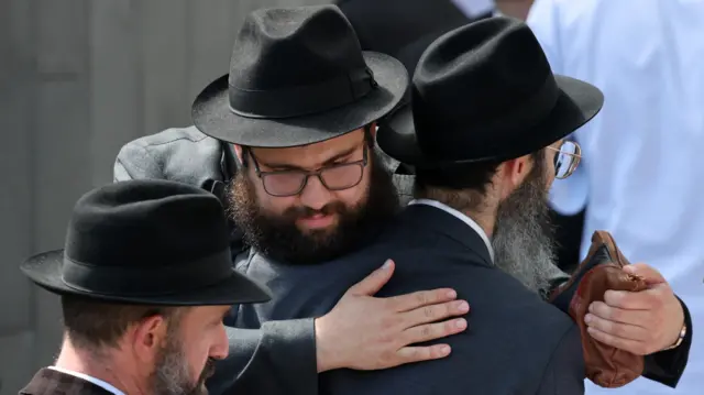 People embrace outside the funeral of rabbi Eli Schlanger, dressed in black.