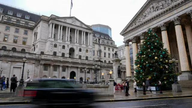 A blurry black cab zooms past the grand Bank of England Building, and a large Christmas tree decorated with gold and green baubles.