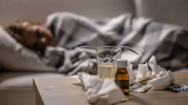 Woman on a sofa in a blanket while assorted medicines on the coffee table in the foreground