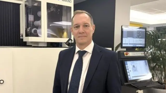 Man in suit stands in office whilst looking into camera
