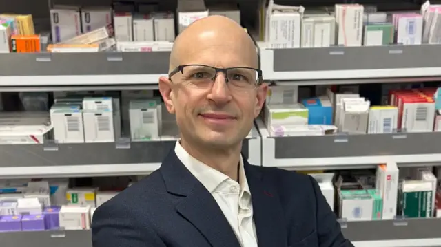 A bald man in glasses and a suit smiles in front of pharmacy stock